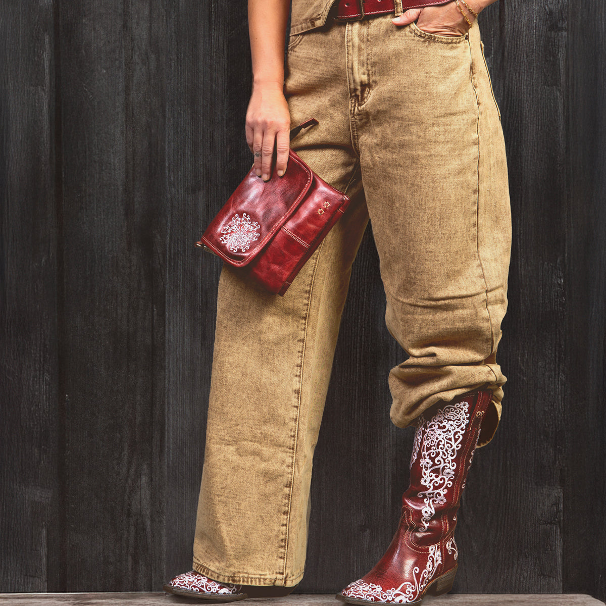 Wearing the Ziggy JB tan organic cotton overalls by Bed|Stü, styled with a red belt, a red clutch, and red cowboy boots with white embroidery, this person stands against a dark wooden wall.