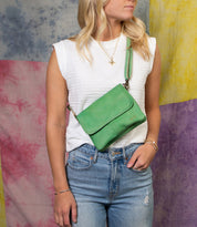 A woman wearing a white sleeveless top and blue jeans poses with the Ziggy green supple leather crossbody bag by Bed|Stü against a colorful fabric background.