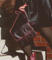 A person in a black leather coat holds the Bed|Stü Ziggy, a dark red crossbody bag with red trim crafted from supple leather, while standing on stairs next to a brick wall.