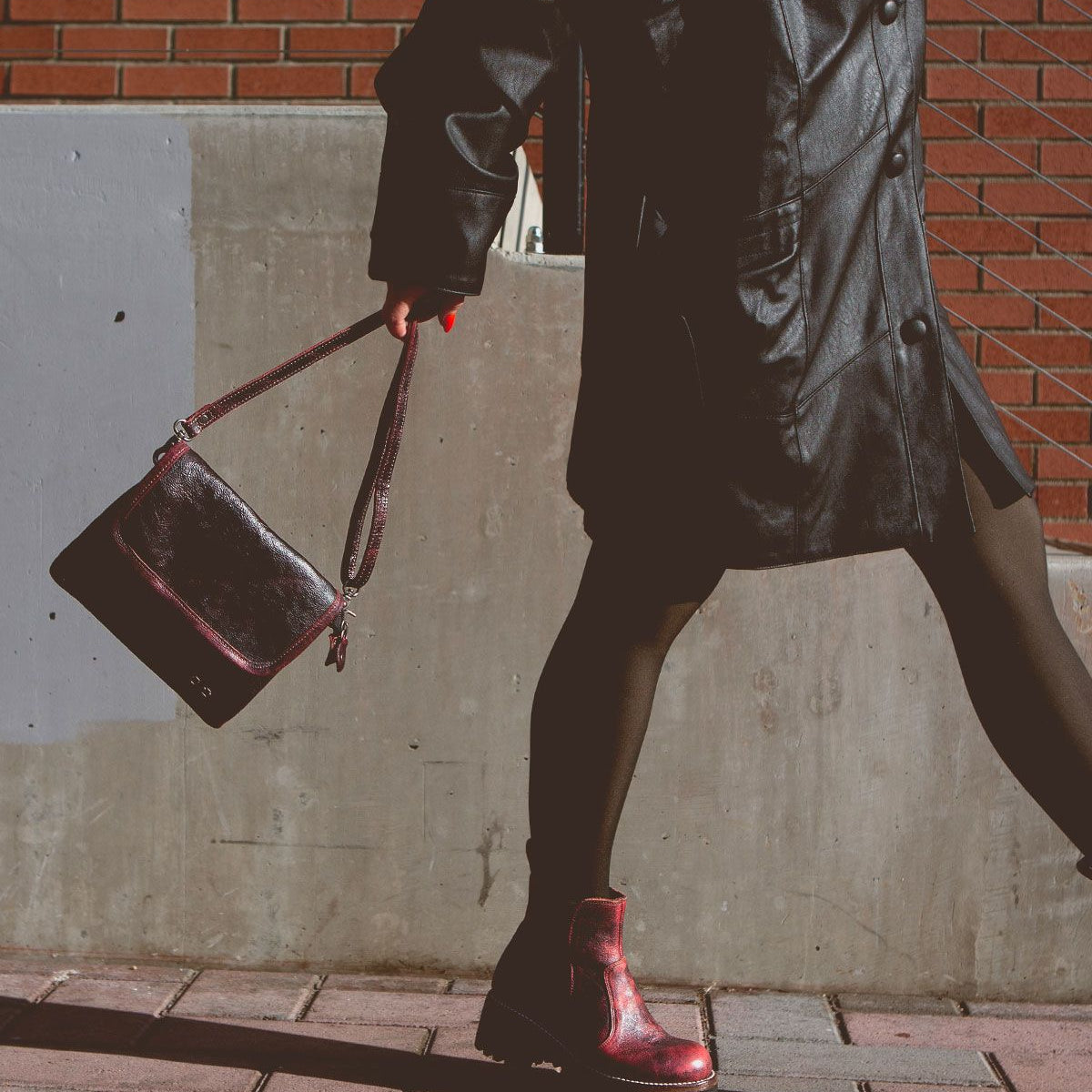 Wearing the Bed|Stü Ziggy black supple leather coat and boots, this person strolls along the sidewalk with a dark belt bag, set against a backdrop of a brick wall and concrete barrier.