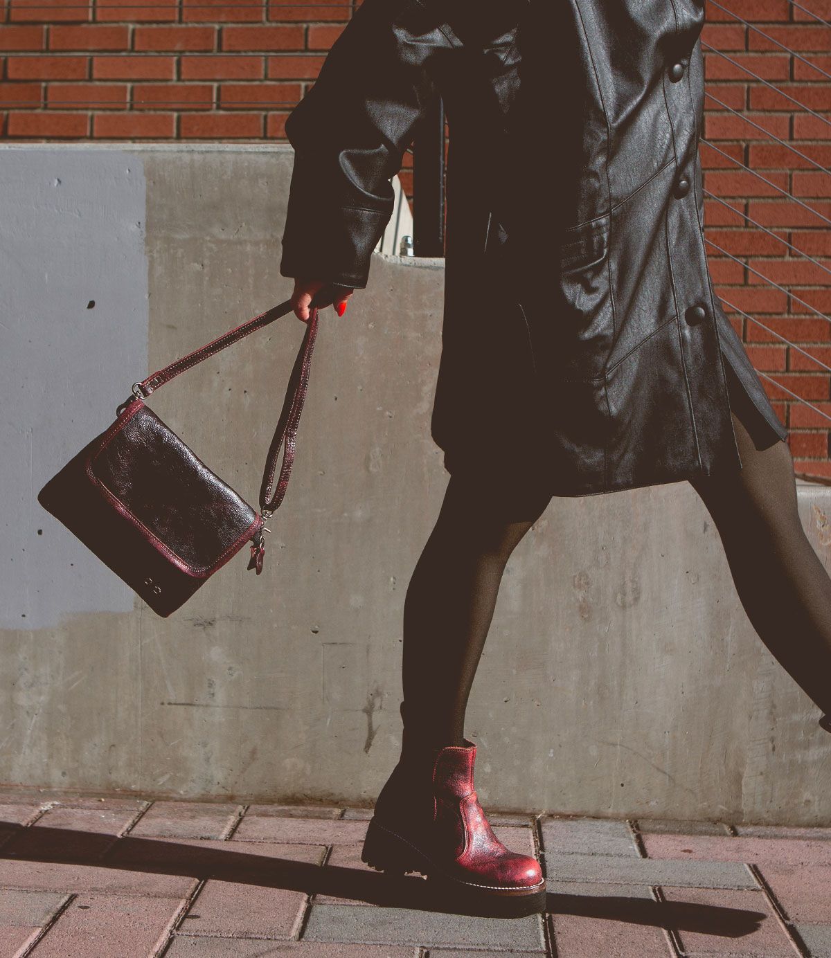 Wearing the Bed|Stü Ziggy black supple leather coat and boots, this person strolls along the sidewalk with a dark belt bag, set against a backdrop of a brick wall and concrete barrier.