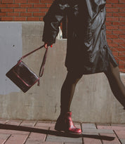 Wearing the Bed|Stü Ziggy black supple leather coat and boots, this person strolls along the sidewalk with a dark belt bag, set against a backdrop of a brick wall and concrete barrier.