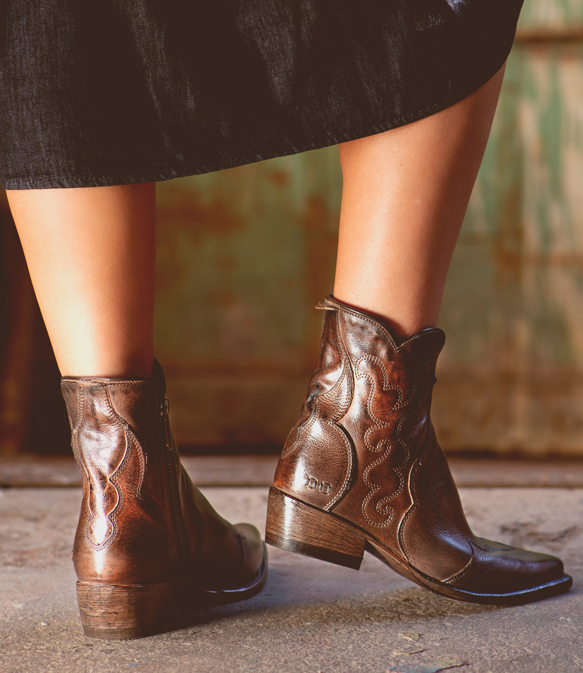 A person wearing a black skirt and Bed|Stü’s Ace brown full-grain leather Western boots stands on a concrete floor with a rustic green and brown background, highlighting the timeless appeal of traditional boot making.