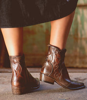 A person wearing a black skirt and Bed|Stü’s Ace brown full-grain leather Western boots stands on a concrete floor with a rustic green and brown background, highlighting the timeless appeal of traditional boot making.