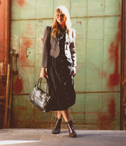 A woman stands in front of a green metal door, wearing a black dress, gray jacket, and Bed|Stü Ace brown Western boots crafted from full-grain leather, holding a black handbag. Sunlight creates shadows on the ground and door.