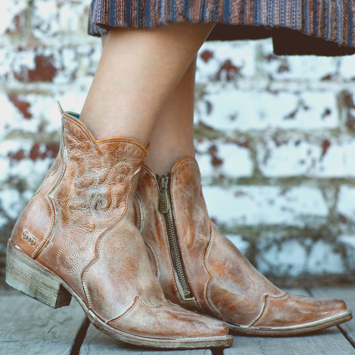 A person wearing the Bed|Stü Ace ankle Western boots in tan full-grain leather with zippers stands on wooden boards. Their skirt features blue, brown, and orange stripes, while a brick wall in the background highlights the traditional craftsmanship of their Bed|Stü footwear.