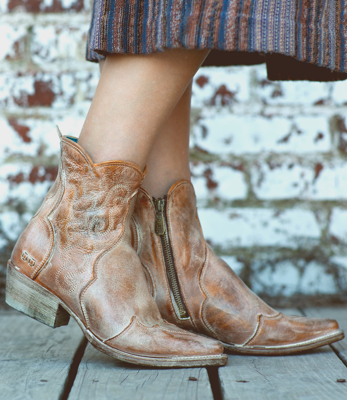 A person wearing the Bed|Stü Ace ankle Western boots in tan full-grain leather with zippers stands on wooden boards. Their skirt features blue, brown, and orange stripes, while a brick wall in the background highlights the traditional craftsmanship of their Bed|Stü footwear.