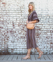 A woman in a striped dress and Western boots walks along a wooden boardwalk in front of a white brick wall, carrying the Bed|Stü "Ace" small woven tan purse.