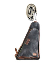 The Bed|Stü Adept is a black and brown compact sling bag crafted from vegetable-tanned leather, featuring a zippered front pocket. It is displayed with its matching adjustable strap rolled up above it.