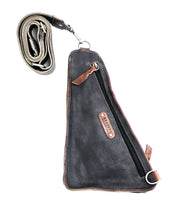 Introducing the Adept by Bed|Stü: a triangular-shaped black compact sling bag, crafted from vegetable-tanned leather. It features a zipper pocket, brown accents, metal rings, and a detachable rolled adjustable strap.