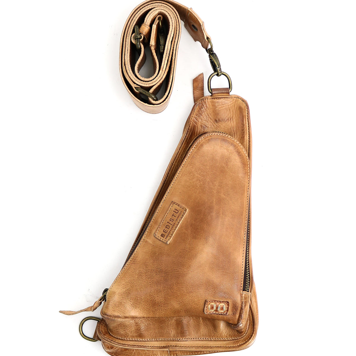 The Adept by Bed|Stü is a compact sling crafted from rich, vegetable-tanned leather, featuring a curved zippered compartment, attached metal hardware, and a rolled, adjustable strap alongside the bag.