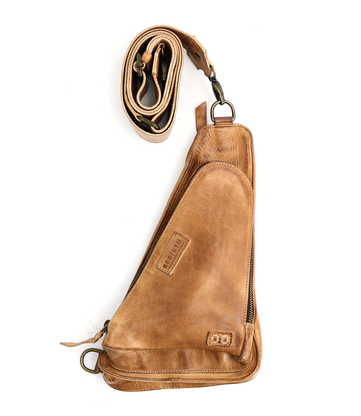 The Adept by Bed|Stü is a compact sling crafted from rich, vegetable-tanned leather, featuring a curved zippered compartment, attached metal hardware, and a rolled, adjustable strap alongside the bag.