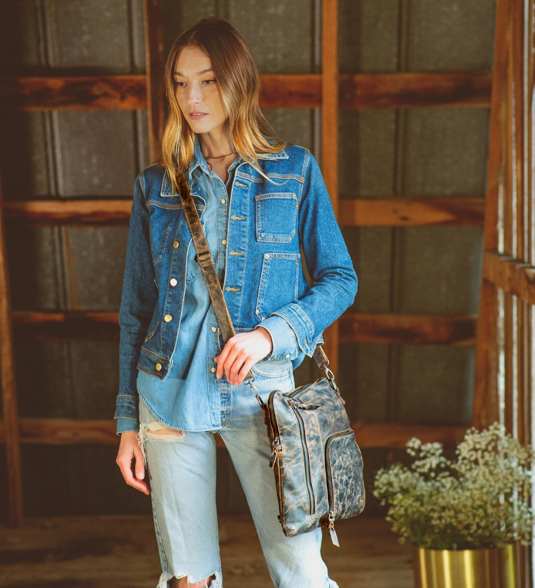 A woman in a denim jacket and ripped jeans stands indoors, holding the Bed|Stü Aiken crossbody bag with an adjustable strap. A potted plant is visible beside her against a wooden wall.