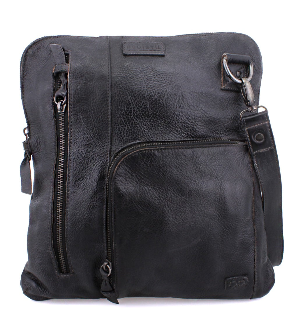 The Bed|Stü Aiken is a black full-grain leather crossbody bag with a textured surface, two zippered pockets on the front, an adjustable strap, and a rectangular silhouette. It features anti-theft security elements and displays a brand patch near the top.