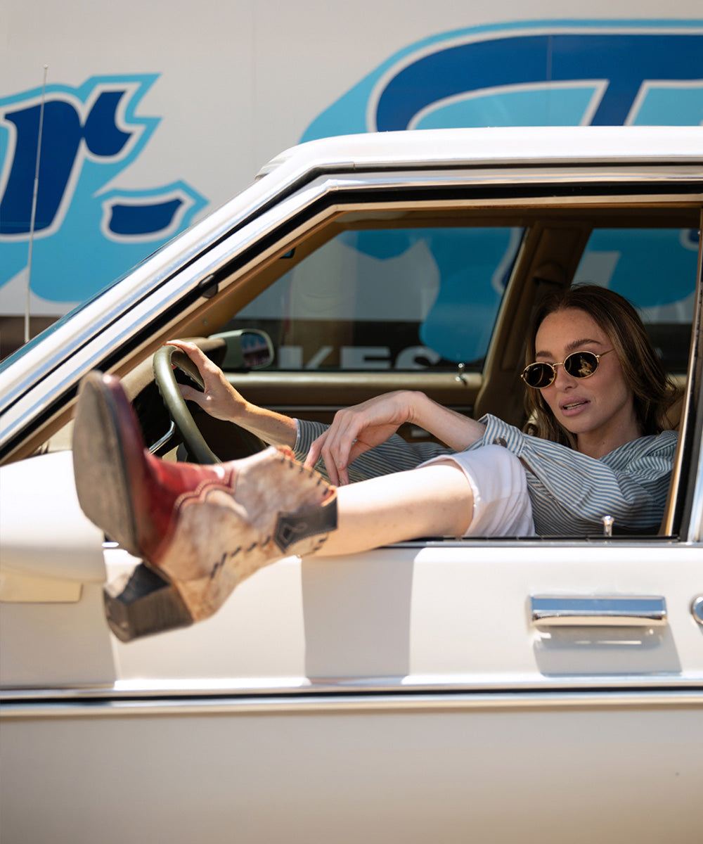 Woman wearing sunglasses sits in a vintage car with her booted feet out the open window, showcasing distressed red-and-white western boots in a sunny retro diner setting.