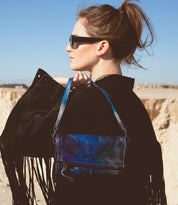 A woman in sunglasses and a black fringed jacket stands on sand, holding the Bed|Stü Amina dark blue iridescent crossbody handbag over her shoulder, looking to the side.