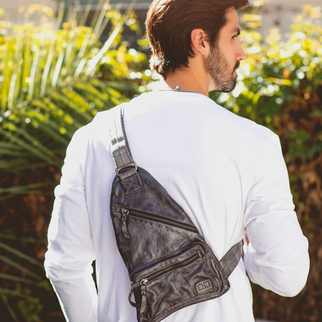 A man wearing a white long-sleeve shirt stands outdoors with the Bed|Stü Andie black leather sling bag, featuring an adjustable strap, slung across his back.