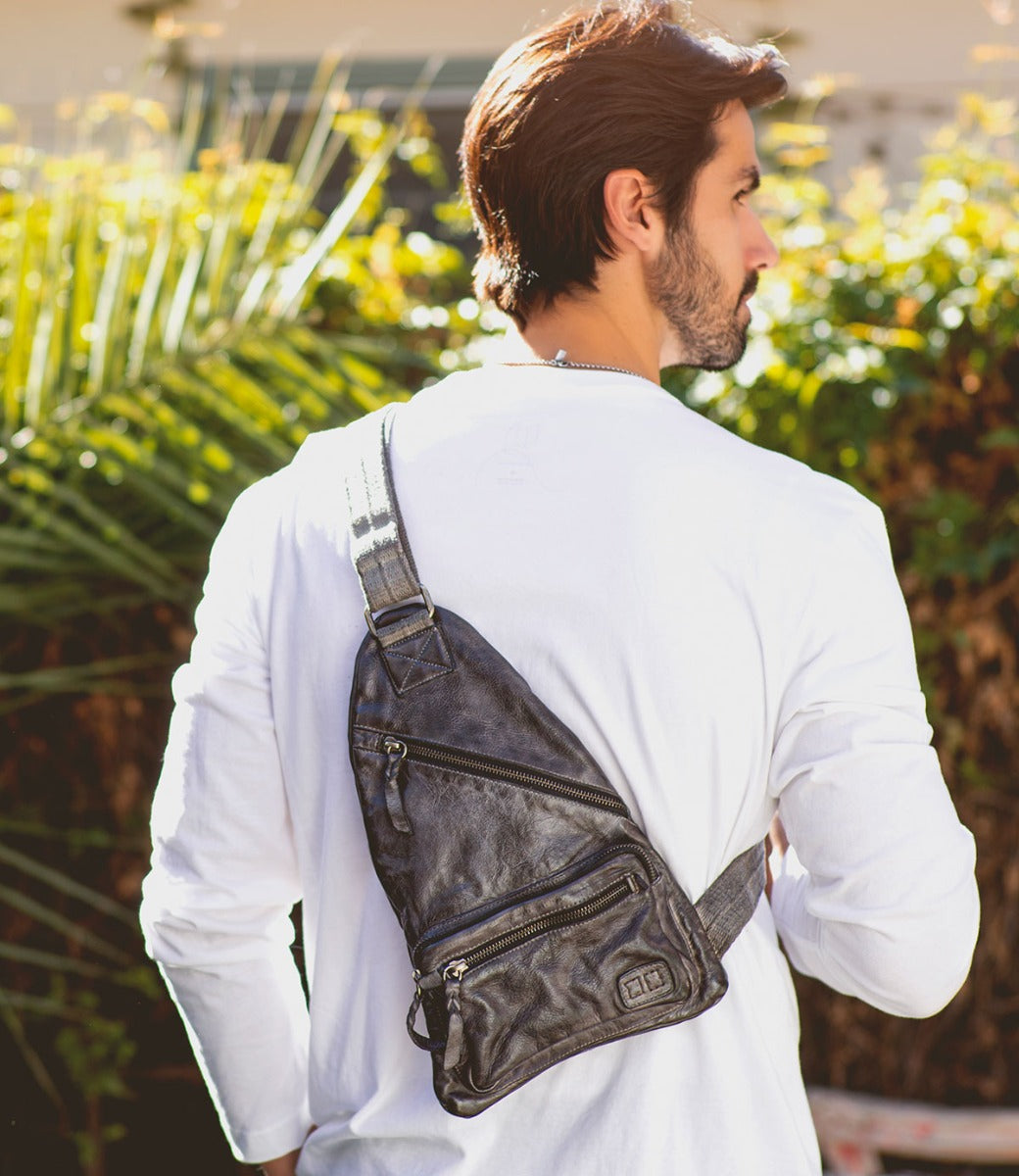 A man wearing a white long-sleeve shirt stands outdoors with the Bed|Stü Andie black leather sling bag, featuring an adjustable strap, slung across his back.