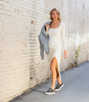 A woman walks along an alley with a light-colored brick wall, wearing a cream sweater dress and Bed|Stü Azeli black sneakers—designed with a durable rubber outsole for all-day comfort—and holds a gray jacket over her shoulder.