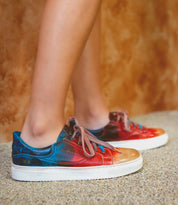 A person wearing Bed|Stü Azeli multicolored leather sneakers featuring blue, red, and orange hues stands on a speckled floor against a brown textured wall, enjoying all-day comfort supported by a durable rubber outsole.