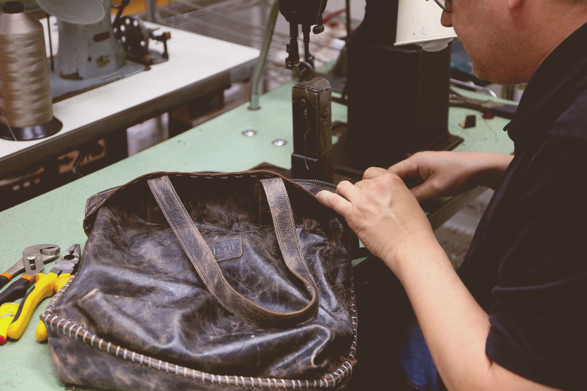 Craftsperson sewing or repairing a dark leather bag at a workshop table.