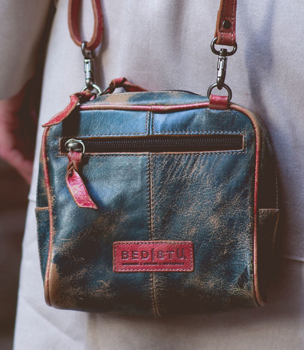 Worn by someone in a beige outfit, the handcrafted "Capture" crossbody bag by Bed|Stü features a small, square silhouette in distressed leather, a front zipper pocket, and a signature red "BED|STU" label.