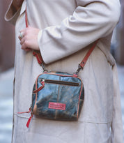 A person wearing a light beige coat is carrying the Bed|Stü Capture, a small, handcrafted leather crossbody bag featuring a worn finish, red zipper and strap, and a visible Bed|Stü patch on the front.