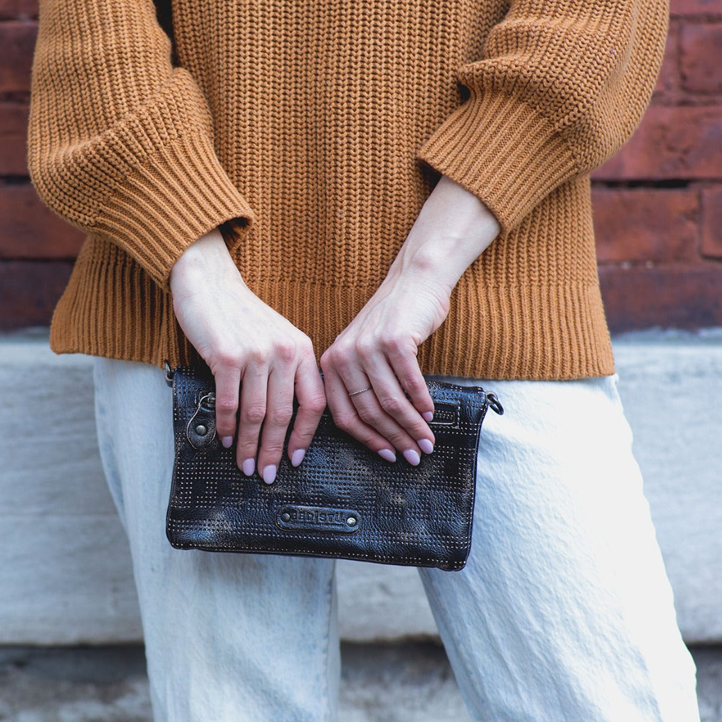 Wearing a brown knit sweater and white pants, the person holds the Bed|Stü Bayshore – a small black textured versatile clutch bag – with both hands while standing against a brick wall.