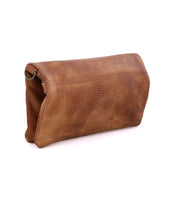 Introducing the Bayshore by Bed|Stü: a brown leather clutch purse featuring a fold-over flap, subtle perforated detailing, and a metal ring accent on one side, displayed against a white background. This versatile clutch effortlessly transitions from day to night and can also be used as a sleek travel wallet for your essentials.