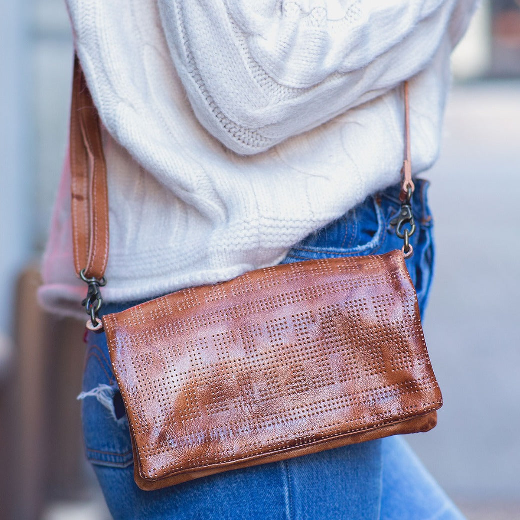 A person wearing a white sweater and blue jeans carries the Bed|Stü Bayshore, a versatile crossbody bag with a perforated pattern that also doubles as a convenient travel wallet.
