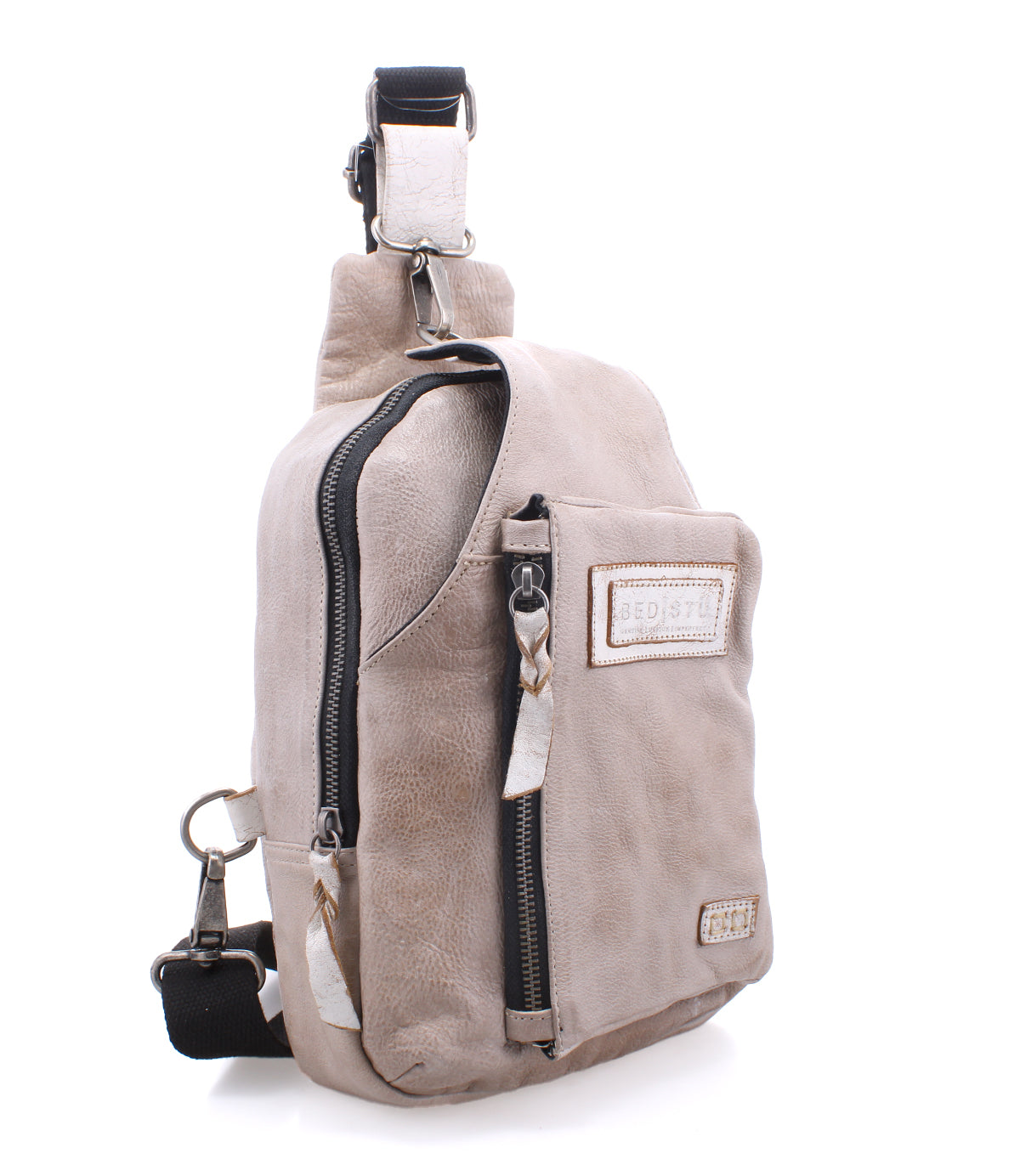 The "Beau" sling backpack by Bed|Stü features light beige leather, a black adjustable strap, a front zipper pocket, and a stitched patch on the front, shown against a white background.