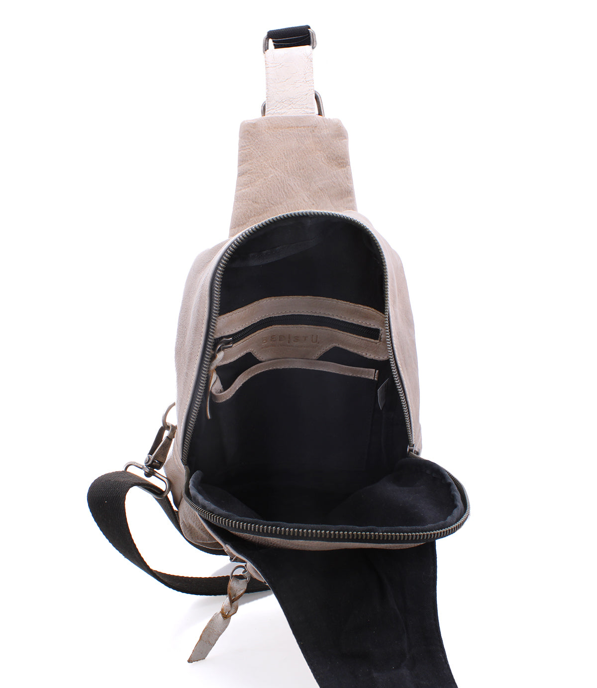 The Beau by Bed|Stü is a beige leather sling bag with a black interior, shown open to reveal both zippered and slip pockets. This compact backpack features a single adjustable strap and silver zipper hardware.