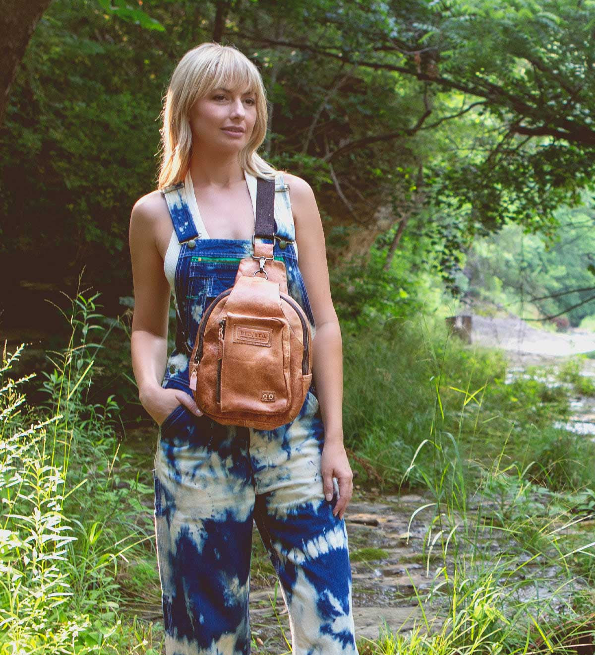 A woman wearing the Beau tie-dye overalls by Bed|Stü and carrying a compact backpack stands outdoors in a lush, green, wooded area near a creek.