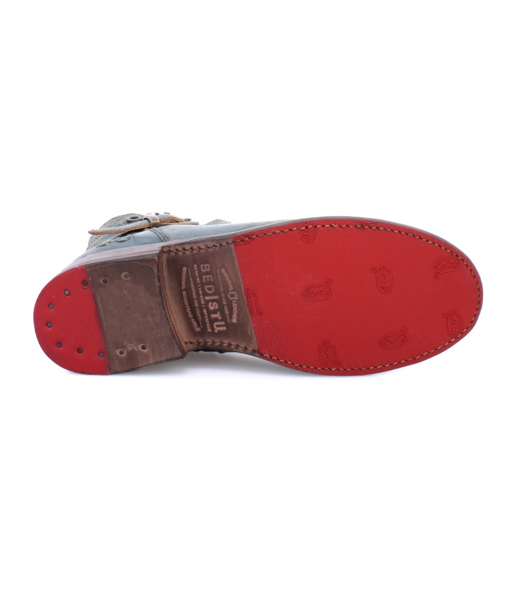 Bottom view of the Bed|Stü "Becca" shoe featuring a red sole, visible stitching, and "Bed|Stü" branding on the arch. The heel is finished with small metal rivets, while the leather shaft displays engineer boot-inspired details including an ankle buckle accent.