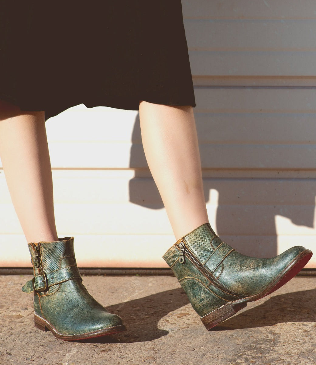 A person wearing the Becca green ankle boots with leather shafts and ankle buckles from Bed|Stü, paired with a black skirt, stands on pavement in sunlight.