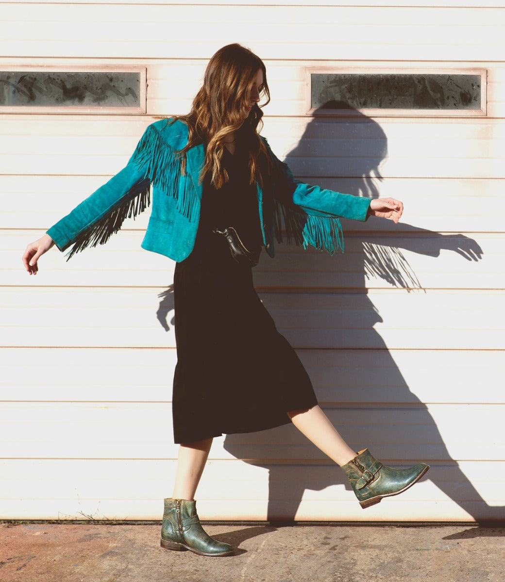 A woman in a turquoise fringed jacket, black dress, and striking green Bed|Stü Becca engineer boots with an ankle buckle walks in front of a white garage door, casting a shadow on the wall.