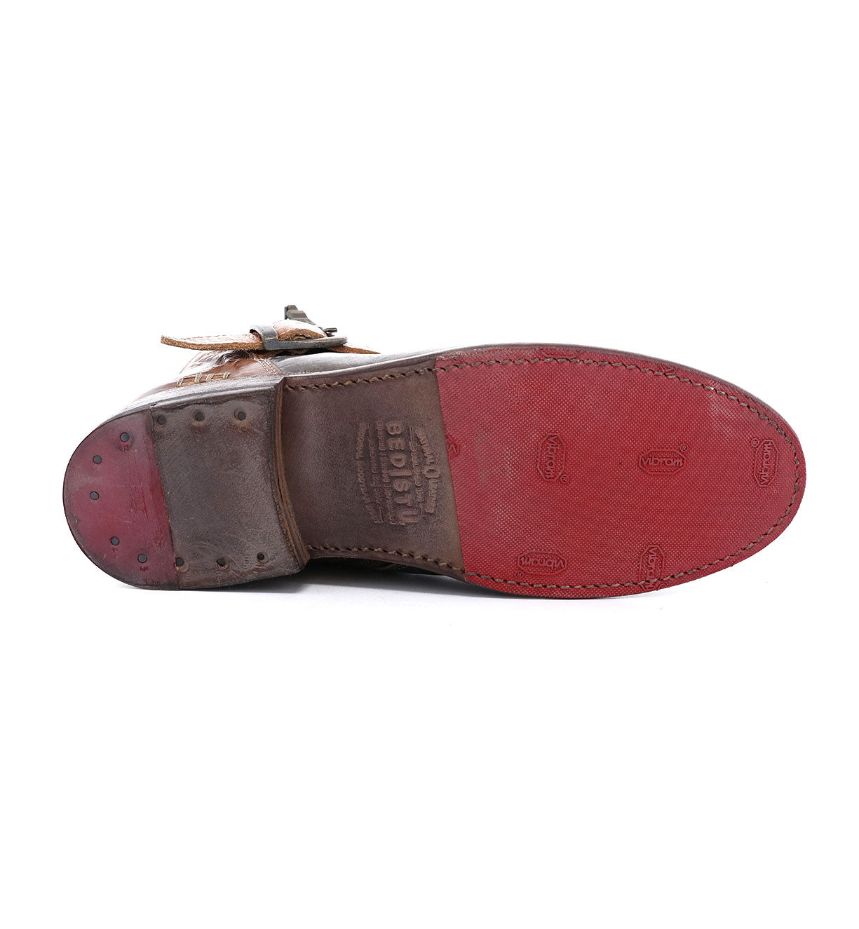 The image features the sole of the Becca by Bed|Stü, a brown leather dress shoe distinguished by its red rubber section and visible stitching, displayed sole up and showcasing durable craftsmanship similar to that of an engineer boot with a sturdy leather shaft.