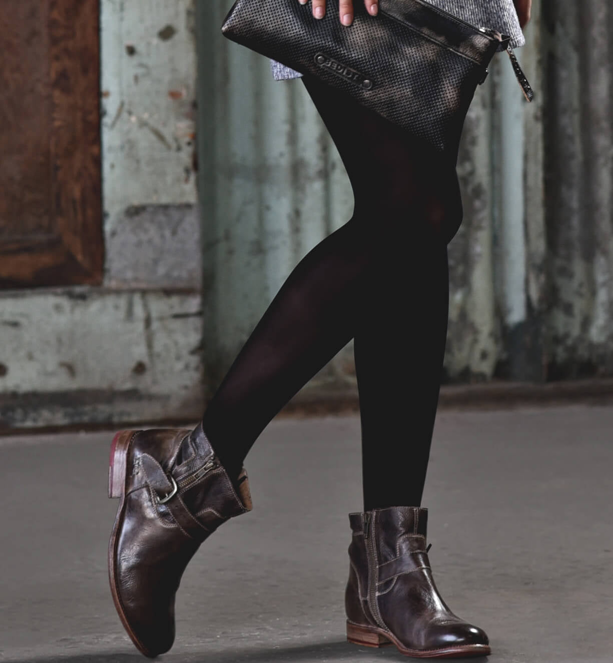Wearing black tights and Bed|Stü’s Becca brown leather ankle boots with an ankle buckle and soft leather shaft, and holding a textured black clutch bag, the person walks indoors on a concrete floor.
