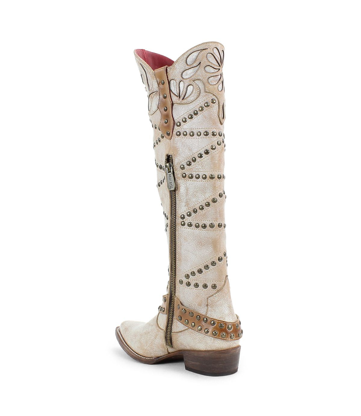 The Betting by Bed|Stü is a tall, light beige handcrafted leather cowboy boot featuring studded swirl designs, a side zipper, brown heel, pointed toe, and subtle studded harness accents for added style.