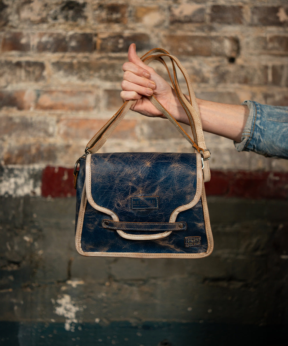 Hand holding a distressed navy leather crossbody bag with cream trim and straps against a weathered brick wall.