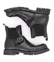 The Bed|Stü Boast is a pair of men's Chelsea boots, expertly crafted from black full-grain leather with chunky soles, side elastic panels, and a distinctive decorative buckle. One boot stands upright while the other lies flat beneath it, highlighting their stylish design and robust construction.