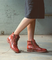 A person wearing a dark skirt and the Bed|Stü Bonnie II brown combat ankle boots with side zippers and cap toe stands on a concrete floor; one boot’s lace is untied.