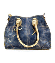 The Bruna bag by Bed|Stü is a blue, distressed two-toned leather handbag featuring beige handles and corner patches, displayed against a white background.