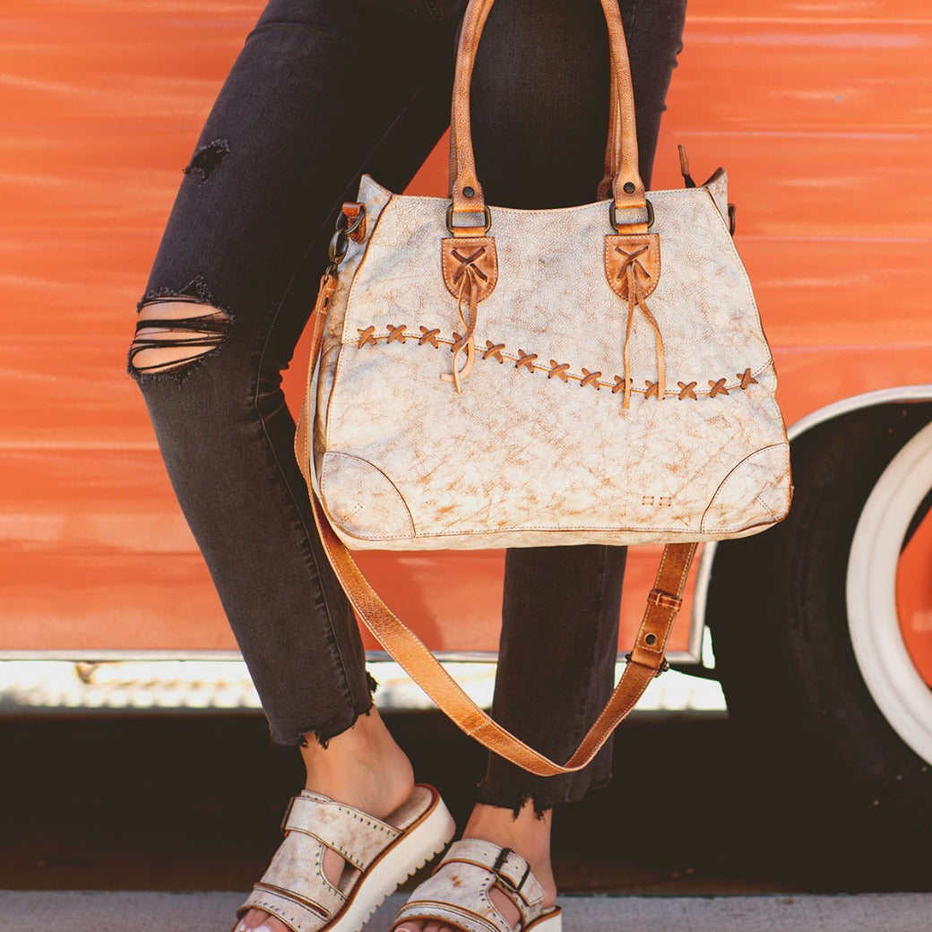 Person in ripped black jeans and beige sandals holding the Bruna two-toned leather bag with padded straps from Bed|Stü, standing in front of an orange vehicle with a white tire.