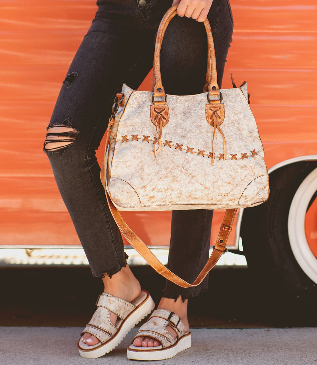 Person in ripped black jeans and beige sandals holding the Bruna two-toned leather bag with padded straps from Bed|Stü, standing in front of an orange vehicle with a white tire.