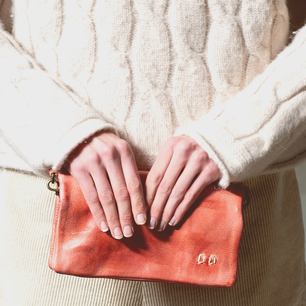 Person wearing a cream cable-knit sweater and corduroy pants holds the Bed|Stü Cadence orange vegetable-tanned leather crossbody clutch with both hands in front of their waist.