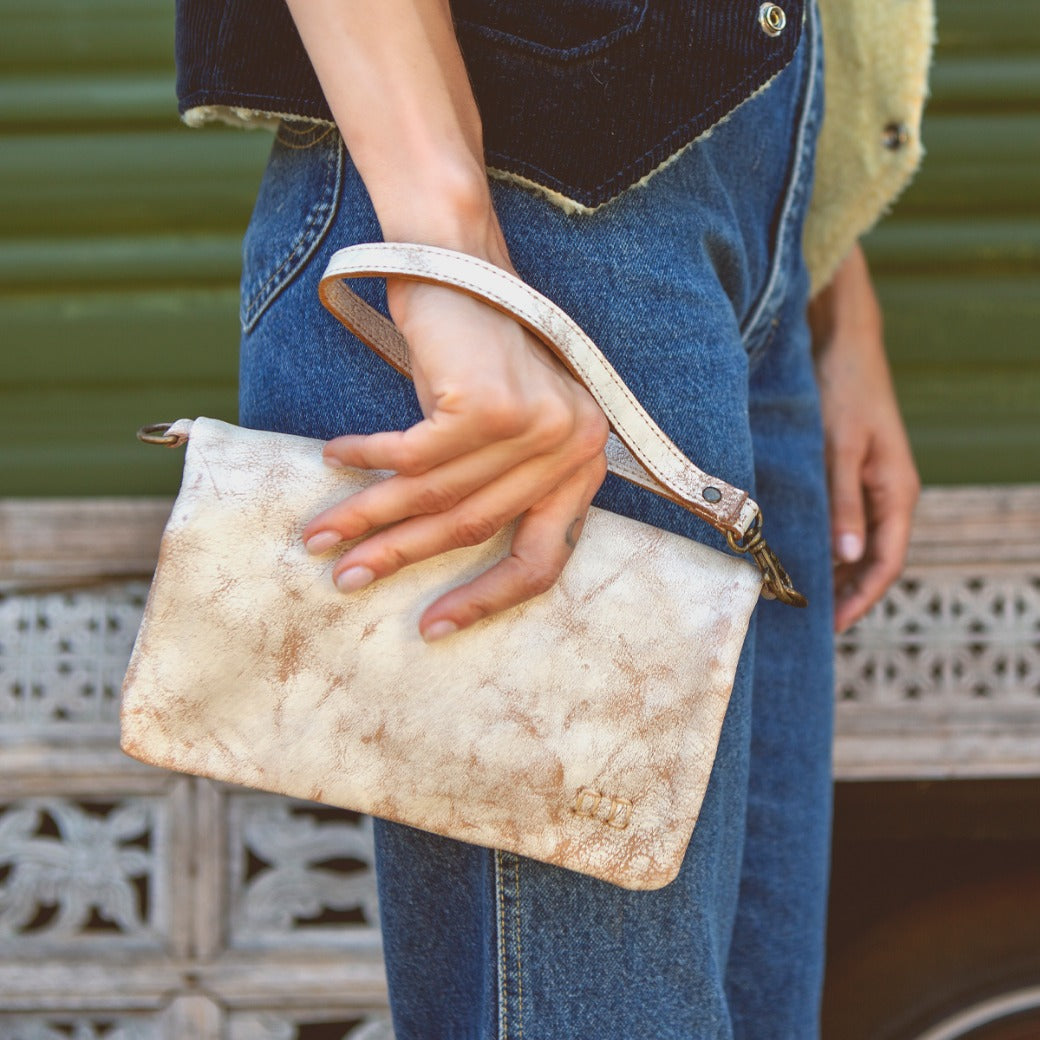 A person wearing jeans and a dark vest holds the Cadence wristlet bag by Bed|Stü—a small, light brown vegetable-tanned leather accessory—by its strap, standing next to a green wall with decorative trim.