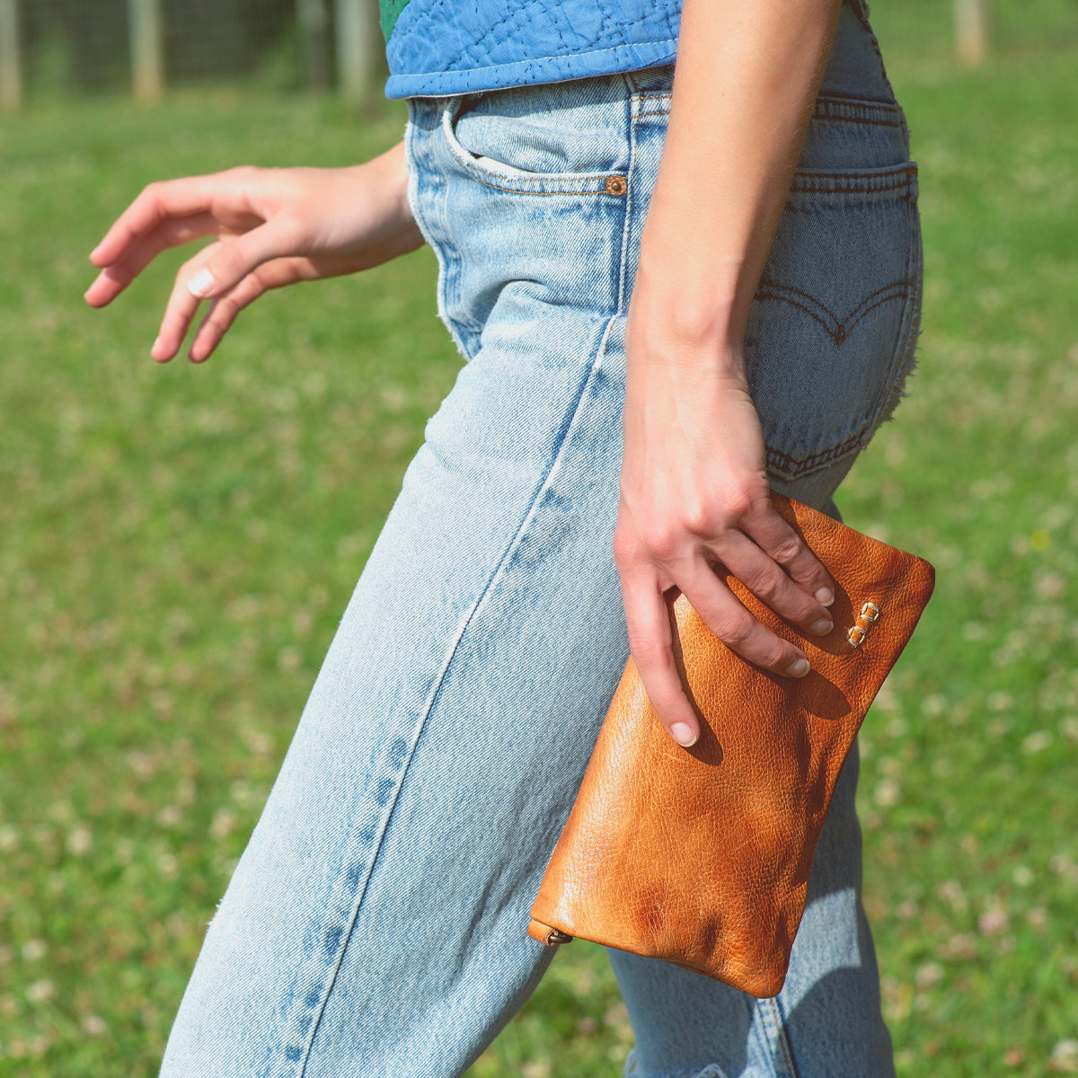 Wearing ripped blue jeans and a green-blue top, the person walks on grass while carrying the Bed|Stü Cadence small brown crossbody clutch.