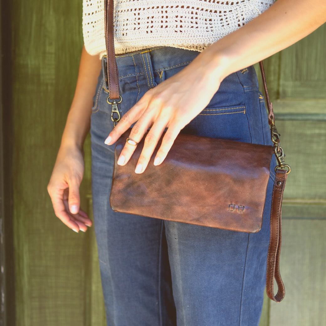 A person dressed in blue jeans and a white crochet top stands in front of a green wooden door, holding the Bed|Stü Cadence 3-in-1 leather wallet with a strap.