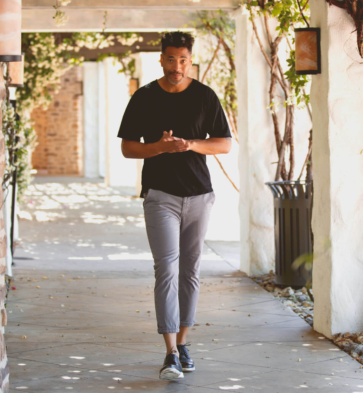 A man wearing a black t-shirt, gray pants, and the Bed|Stü Cayuga II classic derby shoes with subtle brogue detailing walks down a sunlit outdoor corridor lined with vines and plants.